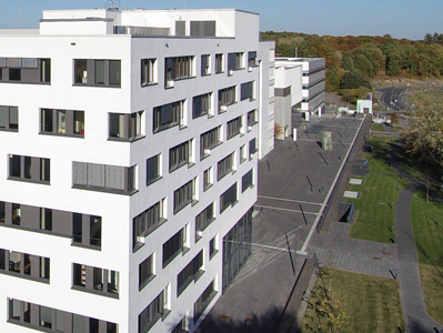 Luftaufnahme eines modernen, weißen Büro- und Verwaltungsgebäudes mit mehreren Stockwerken und vielen rechteckigen Fenstern, umgeben von Grünflächen, Wegen und angrenzender Bebauung.