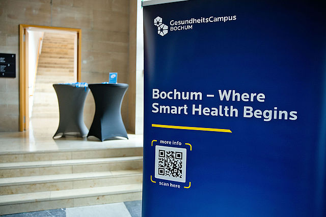 Ein blaues Roll-up-Display im Foyer eines Gebäudes. Es zeigt oben das Logo und den Schriftzug „GesundheitsCampus BOCHUM“. Darunter steht der Slogan „Bochum – Where Smart Health Begins“. Im unteren Bereich befindet sich ein QR-Code mit den Hinweisen „more info“ und „scan here“. Im unscharfen Hintergrund sind Stehtische und Treppenstufen zu sehen