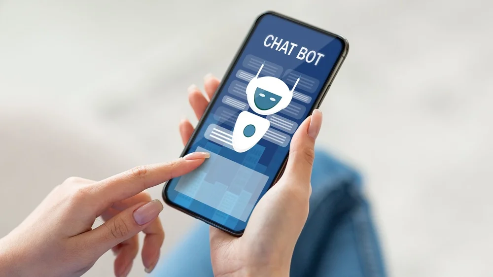 Eine Person hält ein Smartphone in den Händen, auf dessen Display eine Chatbot-Benutzeroberfläche mit stilisiertem Roboter-Avatar und dem Schriftzug „CHAT BOT“ zu sehen ist.