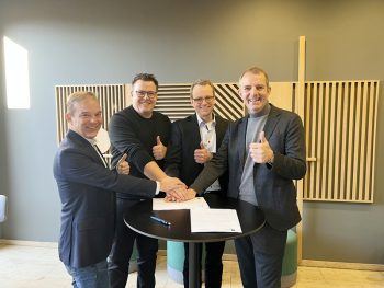 Vier Männer in Businesskleidung stehen um einen runden Stehtisch und geben lächelnd den Daumen nach oben. Auf dem Tisch liegt ein Dokument, das gemeinsam unterzeichnet wird. Die Szene vermittelt eine erfolgreiche Kooperation oder Vertragsunterzeichnung in einem modernen Innenraum.