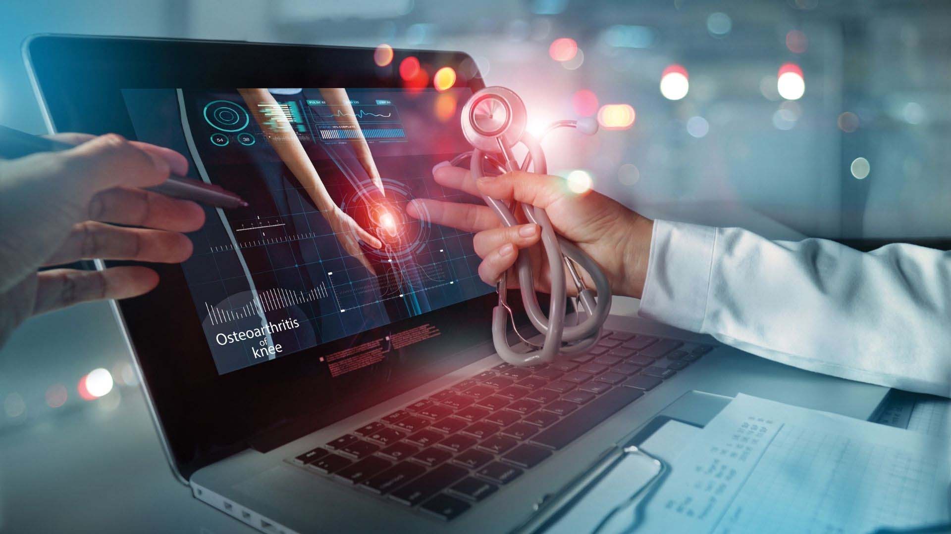 Ärztliche Hände vor einem Laptop mit digitaler medizinischer Visualisierung: Auf dem Bildschirm ist ein Kniegelenk mit hervorgehobener Schmerzstelle und der Aufschrift „Osteoarthritis of knee“ zu sehen. Eine Person zeigt mit einem Stift auf die Darstellung, eine andere hält ein Stethoskop – Symbol für digitale Medizin und Diagnostik.