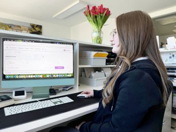 Eine junge Frau mit langen braunen Haaren und Brille sitzt an einem Schreibtisch und arbeitet an einem iMac. Sie schaut auf den Bildschirm, auf dem eine Software-Benutzeroberfläche zu sehen ist, und bedient eine Maus. Im Hintergrund steht eine Vase mit roten Tulpen auf einem Regal.