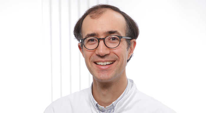 Porträt einer lächelnden Person mit Brille und kurzem, dunklem Haar. Die Person trägt einen weißen Kittel über einem hellen Hemd, was auf einen medizinischen oder wissenschaftlichen Kontext hindeutet. Der Hintergrund ist hell und schlicht gehalten.