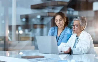 Eine Ärztin und eine Krankenschwester betrachten gemeinsam einen Laptop und besprechen Patienteninformationen in einer modernen medizinischen Einrichtung.