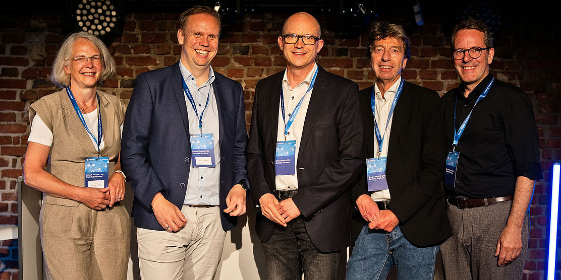 Fünf lächelnde Personen, eine Frau und vier Männer, posieren für ein Gruppenfoto vor einer Ziegelwand. Alle tragen Namensschilder mit dem Text „Smart Health City Summit Bochum“.