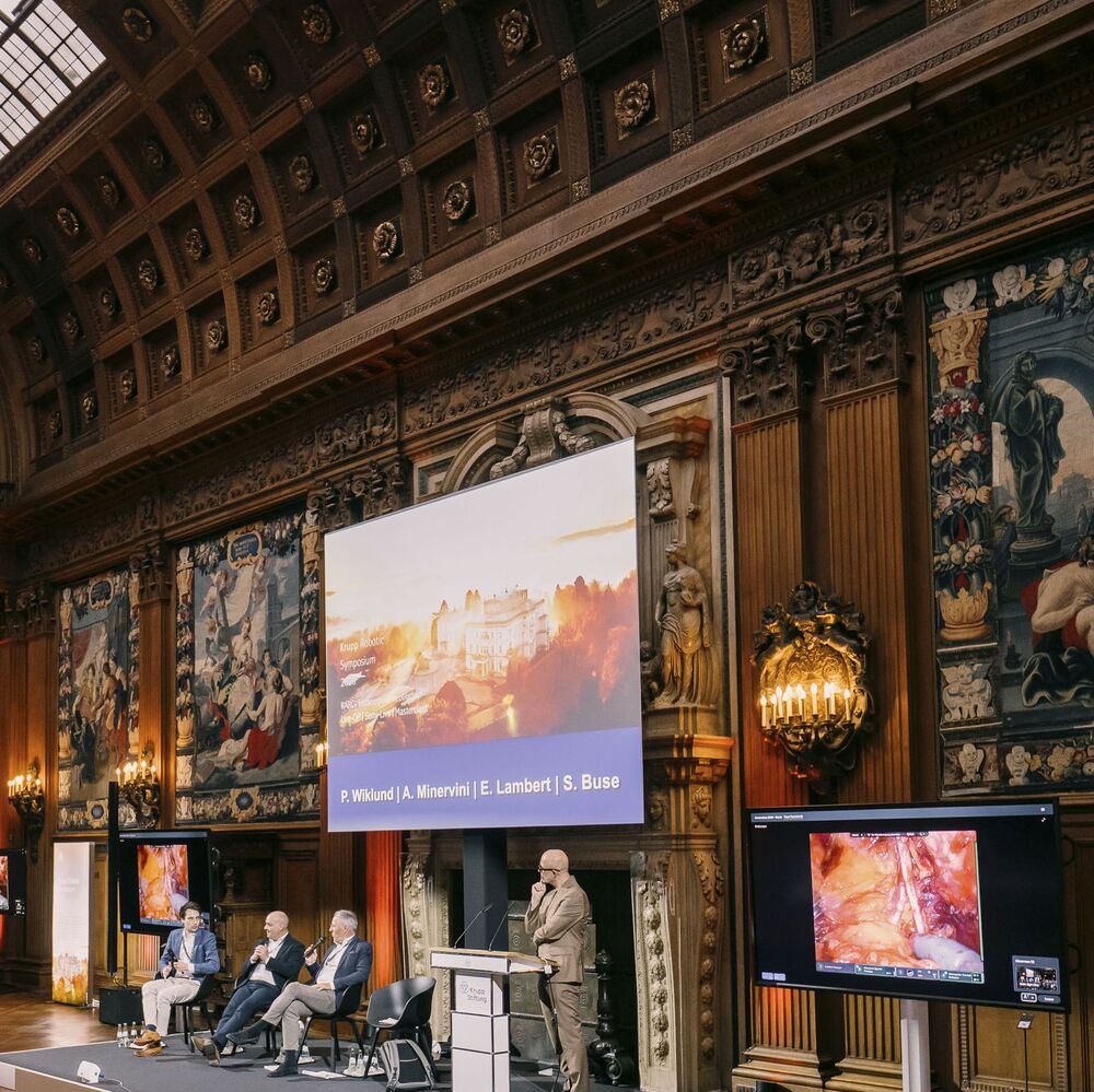 Podiumsdiskussion beim „Krupp Robotic Symposium“ in einem historischen Saal. Ein Moderator steht am Rednerpult, drei Experten sitzen auf der Bühne. Im Hintergrund zeigt eine Leinwand die Namen der Vortragenden (P. Wiklund, A. Minervini, E. Lambert, S. Buse), daneben ein Monitor mit einem medizinischen Endoskopie-Bild.