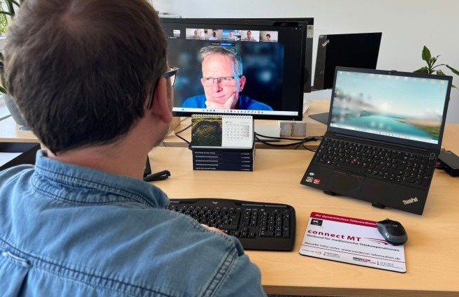 Ein Mann mit Brille und Jeanshemd sitzt an einem Schreibtisch und schaut auf einen großen Computermonitor. Der Monitor zeigt eine Videokonferenz mit einem anderen Mann, der in die Kamera blickt. Rechts steht ein aufgeklappter Laptop auf einem Ständer. Auf dem Schreibtisch liegen eine Tastatur, eine Maus, ein Mauspad mit dem Logo „connect MT“ und ein kleiner Tischkalender. Der Hintergrund ist ein helles Büro mit Pflanzen.