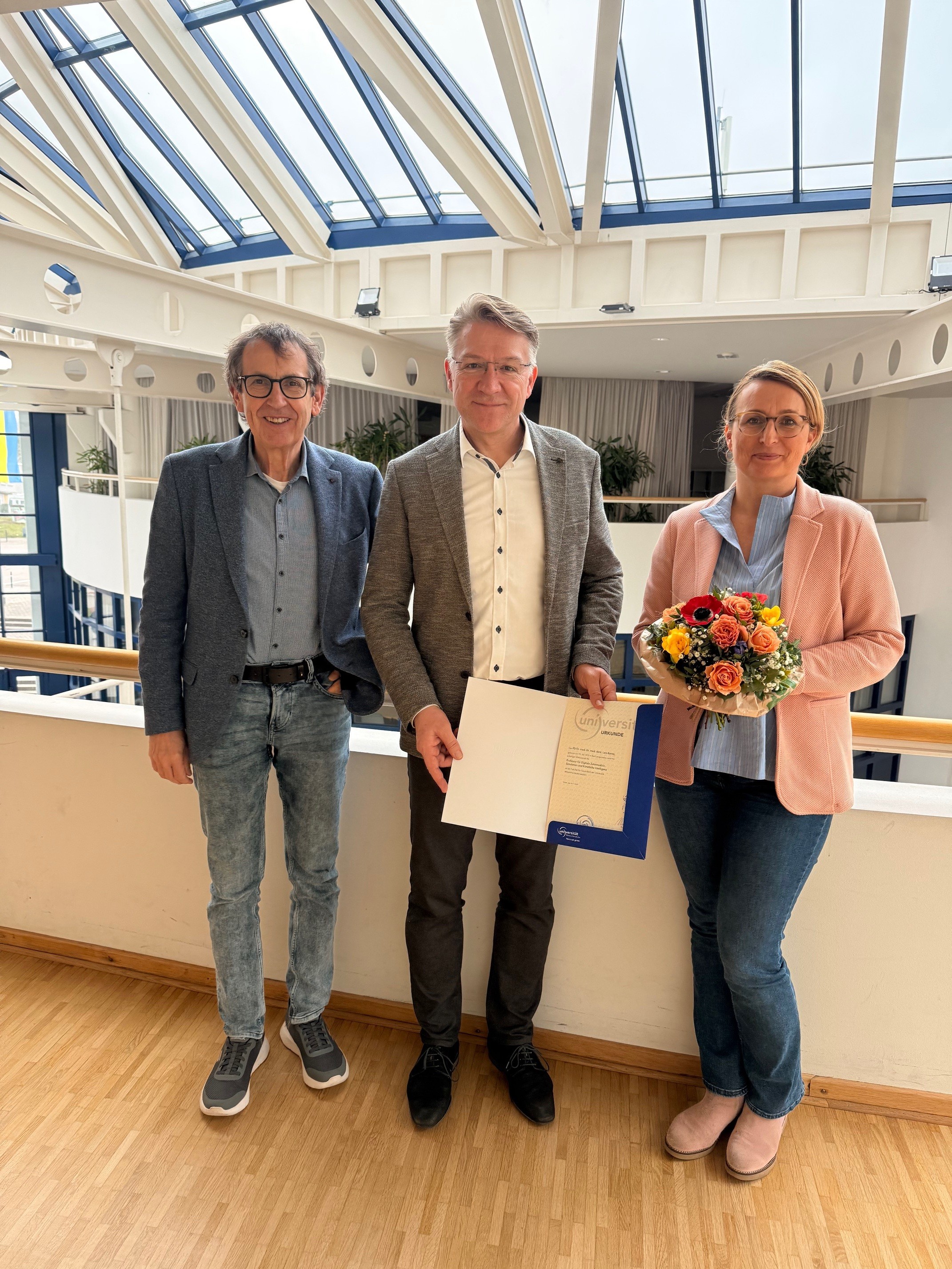 Drei Personen stehen nebeneinander in einem hellen Innenraum mit Glasdach. Der Mann in der Mitte hält eine Urkunde in einer blauen Mappe, die Frau rechts trägt einen bunten Blumenstrauß. Alle drei blicken freundlich in die Kamera. Das Ambiente wirkt wie ein modernes Büro- oder Universitätsgebäude.
